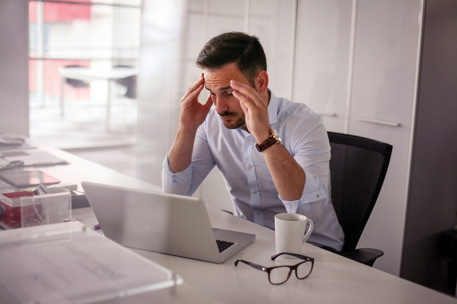 Man at desk holding his head, experiencing a headache while working on a laptop with reading glasses nearby